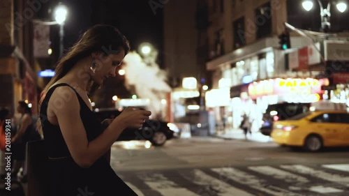 Young beautiful woman standing in downtown, near the crossing road in New York, America and using the smartphone.
