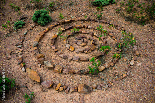 Stone spiral made up of lots of individual rocks on a dry stony floor with green plants growing up through the spiral of stones.