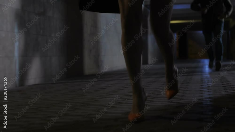 Close-up female legs in high heels running away from male attacker in dark subway passage at night. Woman trying to escape strange man following her. Crimininal approachig his victim. Steadicam