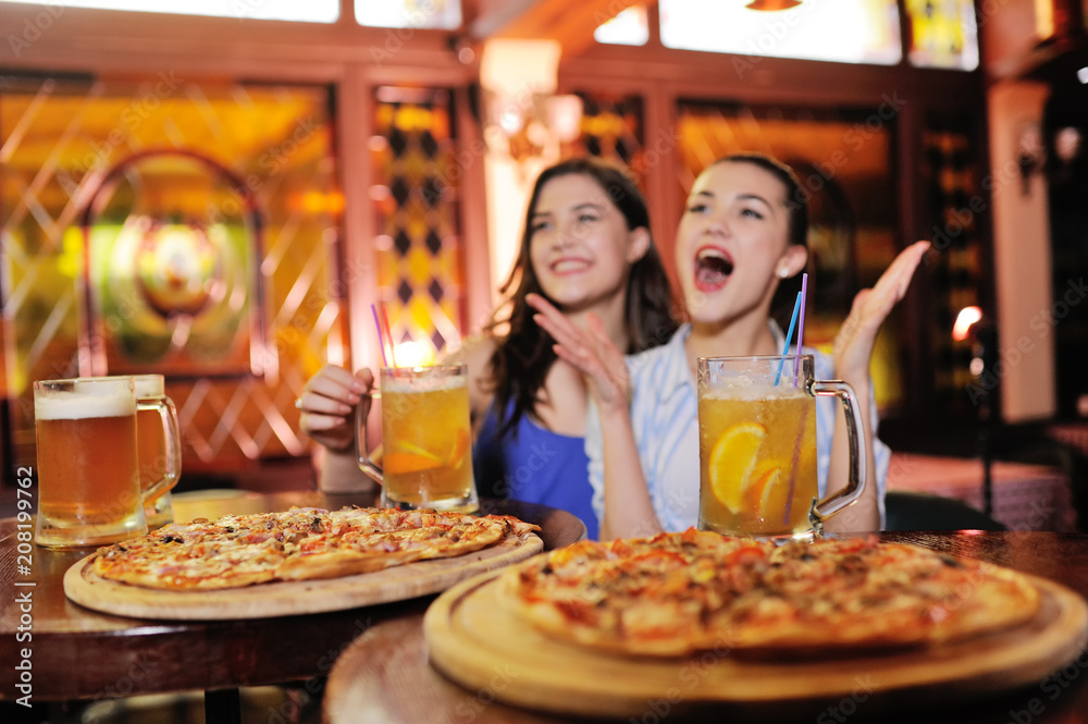 two young pretty girls eating pizza, drinking beer or a beer cocktail and watching football