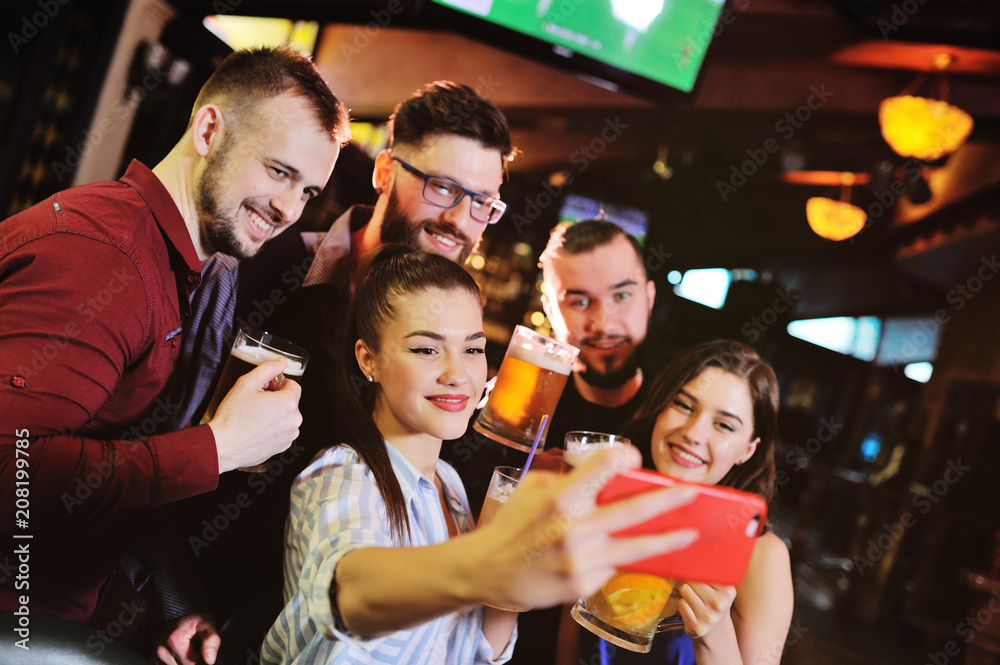 Fototapeta premium group or company of young people - friends drink beer, eat pizza, talk and laugh and shoot selfie on the smartphone's camera on the background of the bar