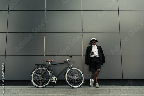 Fashionable african american man wearing fedora hat using smartphone while standing by his bicycle © LIGHTFIELD STUDIOS