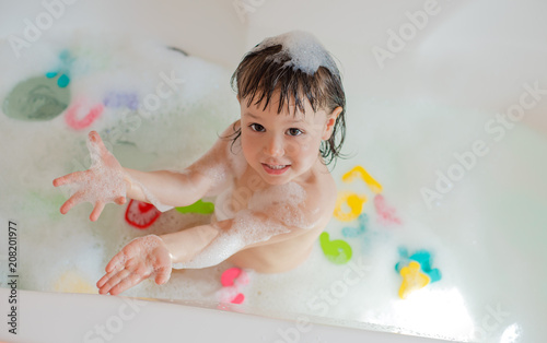 Happy little baby in the bathroom playing with foam bubbles and letters. Infant training and bathing. Hygiene and care for young children.