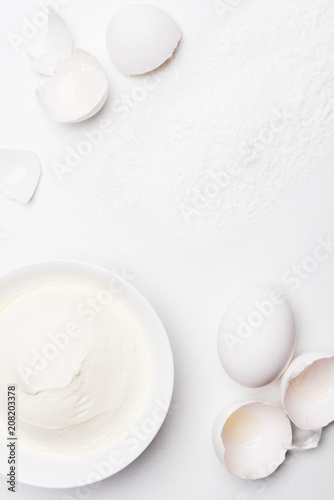 top view of sour cream in bowl and cracked egg shells on white surface spilled with flour