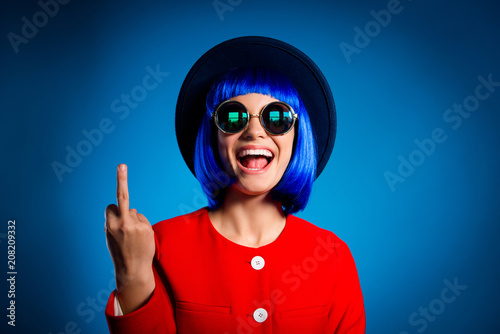 Portrait of cheerful provocative girl in headwear eyewear gesturing fuck off sign with middle finger isolated on blue background. Conflict insult dispute concept