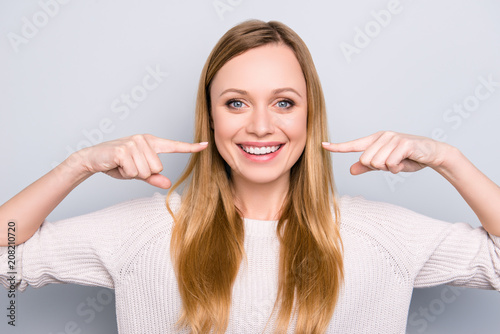 Fototapeta Naklejka Na Ścianę i Meble -  Portrait of joyful satisfied girl gesturing her beaming white healthy teeth with two forefingers looking at camera isolated on grey background. Orthodontic concept