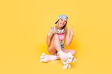 Portrait of active girl with headset on neck gesturing two rock and roll signs with fingers gesturing tongue out sitting on floor ground street style look isolated on yellow background