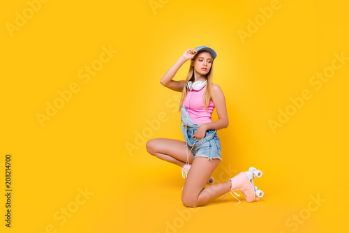 Portrait of pretty charming girl in denim overall with headset on neck holding hand in pocket of shorts looking back posing on one knee isolated on yellow background