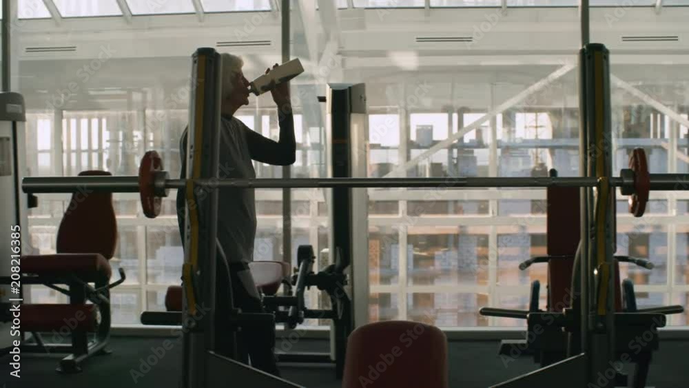 Tracking of tired senior woman with grey hair carrying towel and drinking from sport water bottle while walking in gym after workout Tracking of tired senior woman with grey hair carrying towel and drinking from sport water bottle while walking in gym after workout