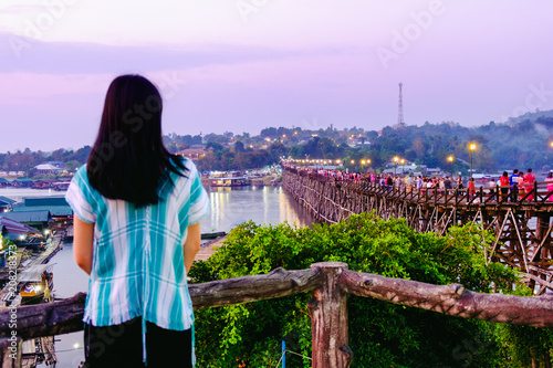 Woman wearing local shirt standing in front of longest wooden Mon Bridge (saphan mon,Uttama Nusorn ) at Sangklaburi Kanchanaburi, Thailand at Sunrise. Beautiful violet sky with Mon Village.