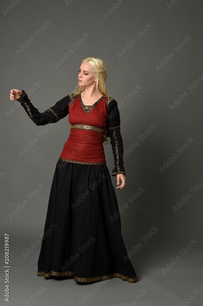 full length portrait of pretty blonde lady wearing  a red and black fantasy medieval gown. standing pose on grey background.
