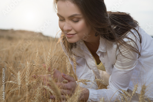 Portrait of serene young woman enjoying scent of ripe golden wheat. She is touching spicas with pleasure. Her eyes are closed  photo