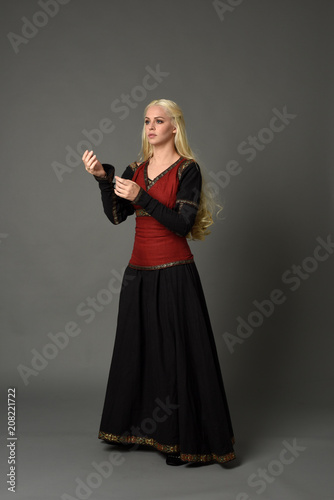 full length portrait of pretty blonde lady wearing  a red and black fantasy medieval gown. standing pose on grey background.