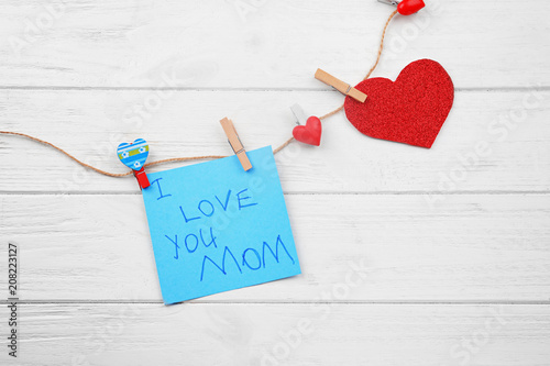 Card with words "I love you Mom", string and pegs on wooden background