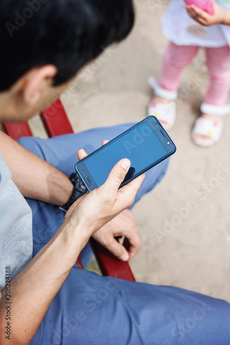 A man sits on the playground and uses a smartphone, does not pay attention to his daughter