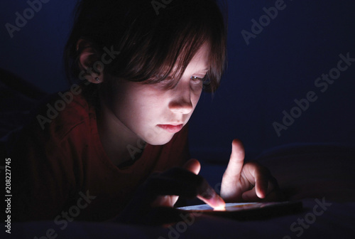 Caucasian girl lying in bed playing tablet in social internet in dark blue light under blacket. Childhood, communication and technology concept
