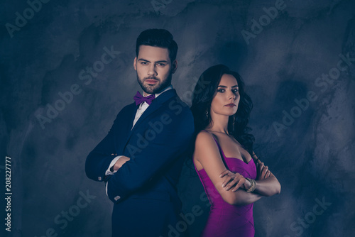 He vs She! Portrait of man in tuxedo and charming pretty brunette woman with curly hair, attractive couple standing back to back with crossed hands looking at camera isolated on grey background