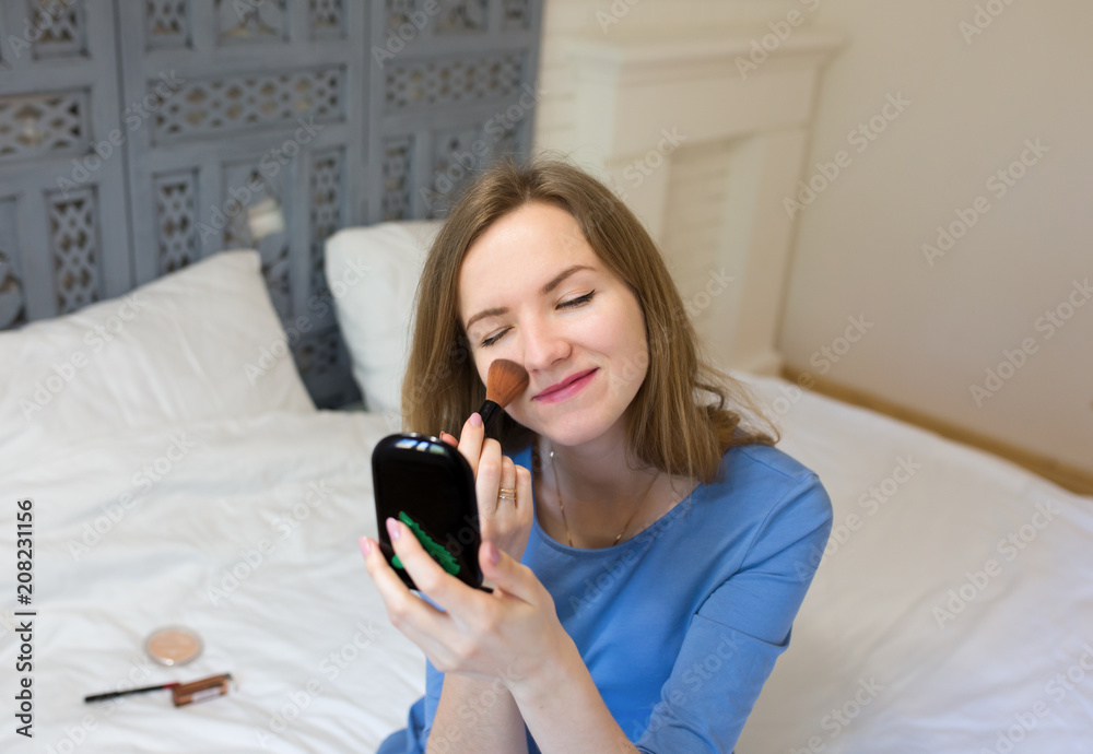 Beautiful girl in blue dress is sitting on white bed linen in minimalist loft design room. Young woman is doing make up. She is looking in the mirror and powders her face. Beautiful girl in blue dress is sitting on white bed linen in minimalist loft design room. Young woman is doing make up. She is looking in the mirror and powders her face.