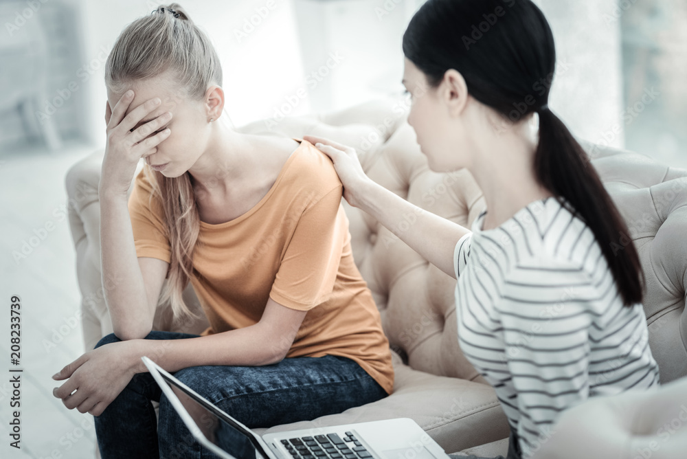 Love problem. Pleasant female psychologist using laptop while posing on sofa and calming down vulnerable teen girl Love problem. Pleasant female psychologist using laptop while posing on sofa and calming down vulnerable teen girl