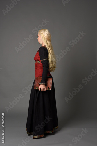  full length portrait of pretty blonde lady wearing  a red and black fantasy medieval gown, holding a book. standing pose on grey background.
