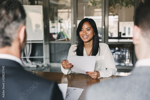 Business, career and placement concept - young asian woman smiling and holding resume, while sitting in front of directors during corporate meeting or job interview