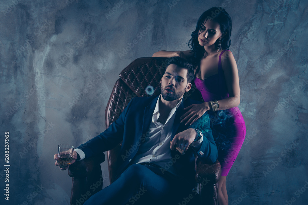 Obraz premium Portrait of virile boss in formalwear sitting in leather chair holding glass with whiskey cigar in hands making circles from fume charming lady standing near isolated on concrete background