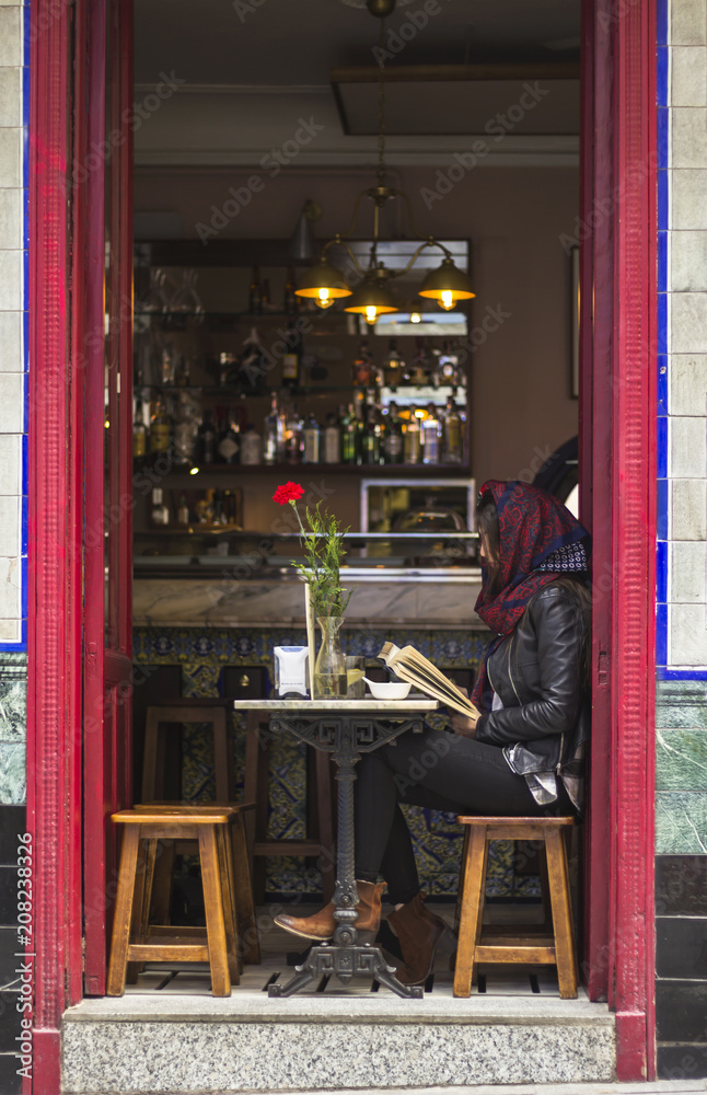 Fototapeta premium Kobieta czytająca książkę w hiszpańskim barze z dużymi otwartymi drzwiami. Miasto Madryt.
