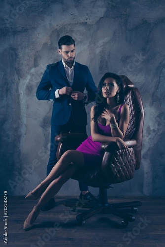 Portrait of sexual hot woman in purple dress sitting in leather chair looking at camera trendy man unbutton jacket looking at bitch having erection couple isolated on grey dark background