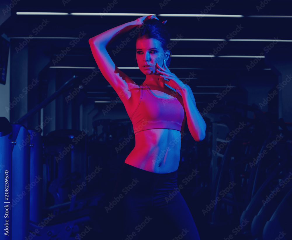 Waist up portrait of young attractive brunette woman dressed in sports clothes illuminated by faint pink light. Gorgeous confident slim female fitness model wearing top and leggings posing at gym. Waist up portrait of young attractive brunette woman dressed in sports clothes illuminated by faint pink light. Gorgeous confident slim female fitness model wearing top and leggings posing at gym.