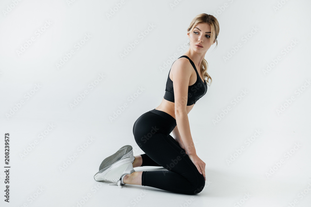 side view of attractive sportswoman in black sportswear sitting on floor isolated on grey