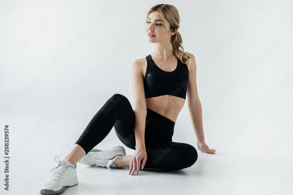 attractive sportswoman in black sportswear sitting on floor isolated on grey