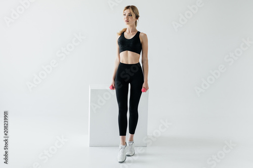 attractive woman in black sportswear with dumbbells in hands and white cube behind on grey backdrop