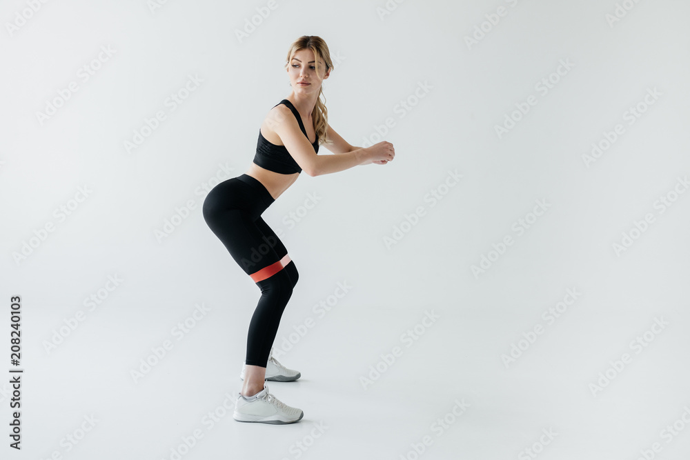 side view of young athletic woman exercising with rubber tape isolated on grey