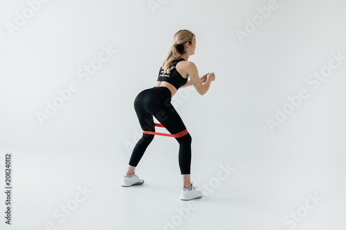 Young athletic sportswoman exercising with rubber tape isolated on grey