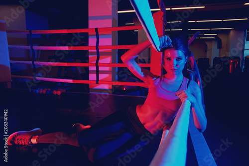 Seductive female fitness model with pony tail hairstyle, dressed in sports bra and leggings, illuminated by dim neon light, lying on left side of her body on boxing ring and leaning on ropes.