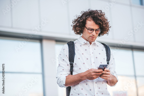 Stylish young serious Caucasian male wearing trendy spectacles and shirt looking at screen of his smart phone, about to answer to some email. People, lifestye and modern technology communication. photo