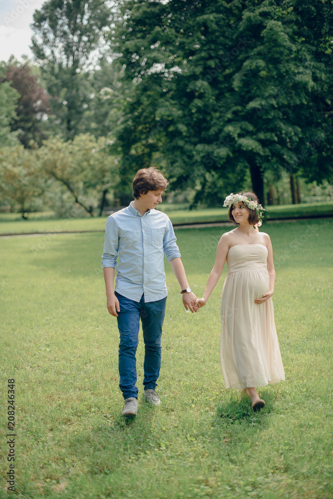 Happy young family of woman and man walking in the green park. Pregnant woman in long dress and flower wreath walking with husband holding hands Happy young family of woman and man walking in the green park. Pregnant woman in long dress and flower wreath walking with husband holding hands