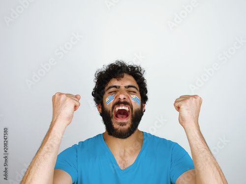 Sport fan screaming celebrating the triumph of his team. Man with the flag of Uruguay makeup on his face and blue t-shirt.                
