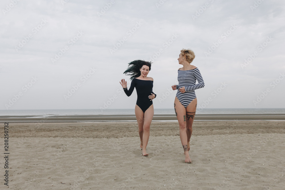 happy young women in bodysuits running by sandy beach on cloudy day happy young women in bodysuits running by sandy beach on cloudy day