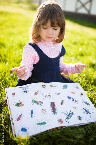 Moscow, Russia. 9 May, 2018. Adorable little girl reading book about bugs, learning nature in the garden.