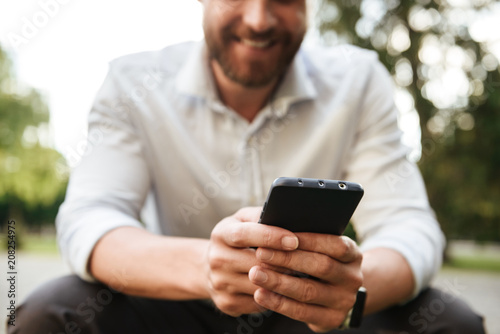 Cropped image of joyous attractive man in white shirt, sitting in park and using black mobile phone for chat