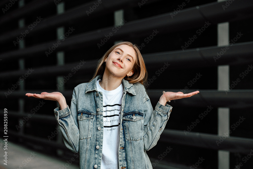 Street style. Portrait of a young student girl with blonde hair without makeup on a gray wall background. girl throws up his hands in bewilderment. she's wearing a denim jacket.