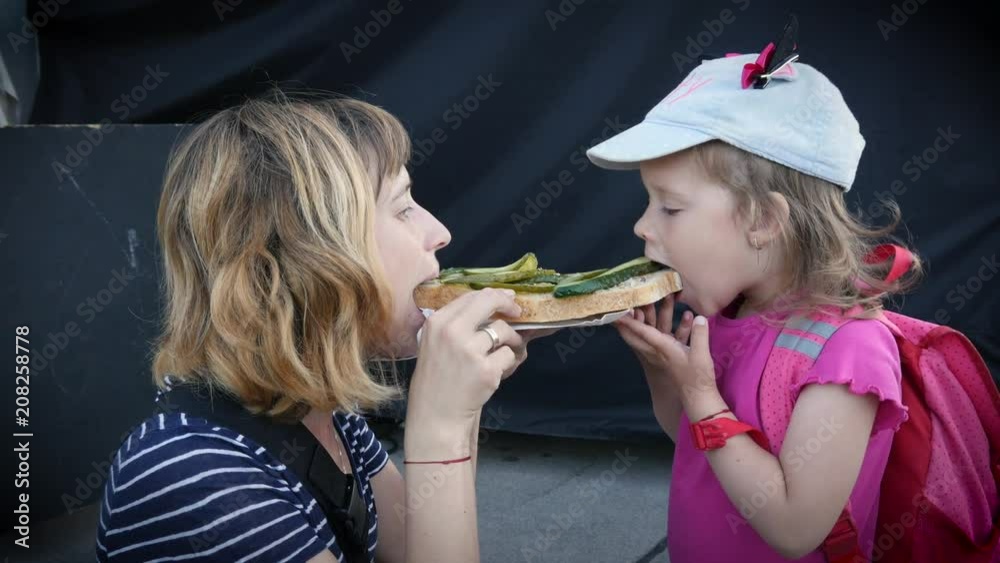 Young mother and little child daughter eating biting a huge sandwich outdoors, family diet Young mother and little child daughter eating biting a huge sandwich outdoors, family diet
