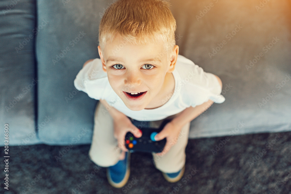 Fototapeta premium Bright buttons. New modern games console in the hands of a pretty little boy sitting on a sofa