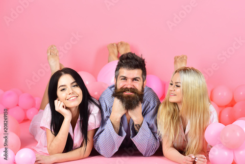 Lovers or best friends in pajamas at girlish bedroom party. Blonde and brunette on smiling faces have fun with bearded macho. Best friends  lovers near balloons  pink background. Relations concept.