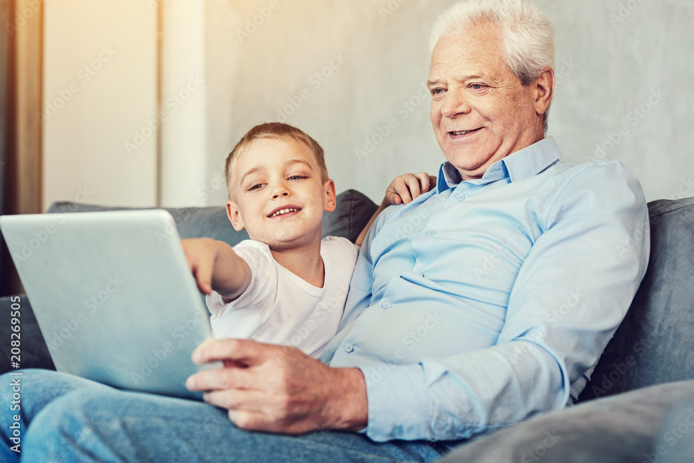 Check it. Cheerful happy little boy pointing at the screen of a big laptop while sitting on a sofa with his pleasant senior grandfather Check it. Cheerful happy little boy pointing at the screen of a big laptop while sitting on a sofa with his pleasant senior grandfather