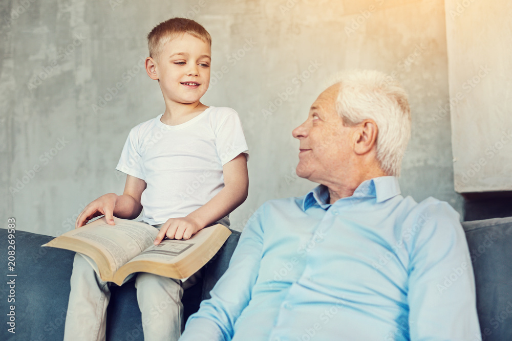 New facts. Curious clever little boy showing a big picture in a book to his attentive loving senior grandfather New facts. Curious clever little boy showing a big picture in a book to his attentive loving senior grandfather