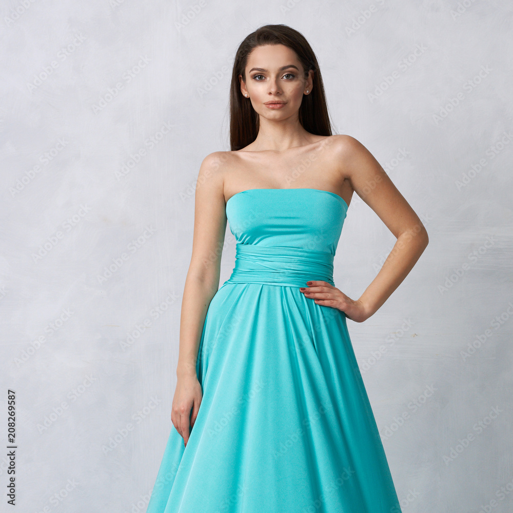 Beautiful long haired young woman dressed in stylish turquoise blue bandeau maxi dress posing against white wall on background. Elegant brunette female model demonstrating evening outfit in studio.