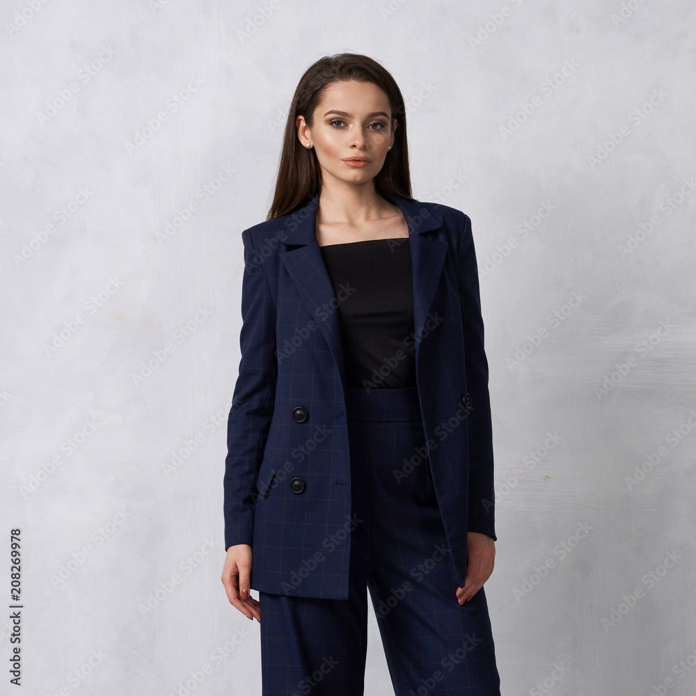 Gorgeous young woman dressed in blue squared jumpsuit, blazer and heeled shoes posing in studio. Beautiful brunette girl demonstrating stylish smart clothing against white wall on background. Gorgeous young woman dressed in blue squared jumpsuit, blazer and heeled shoes posing in studio. Beautiful brunette girl demonstrating stylish smart clothing against white wall on background.