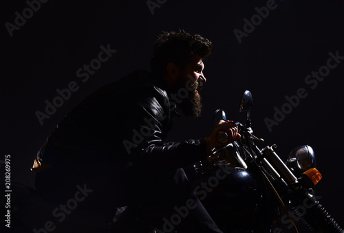 Man with beard, biker in leather jacket sitting on motor bike in darkness, black background. Masculinity concept. Macho, brutal biker in leather jacket riding motorcycle at night time, copy space.